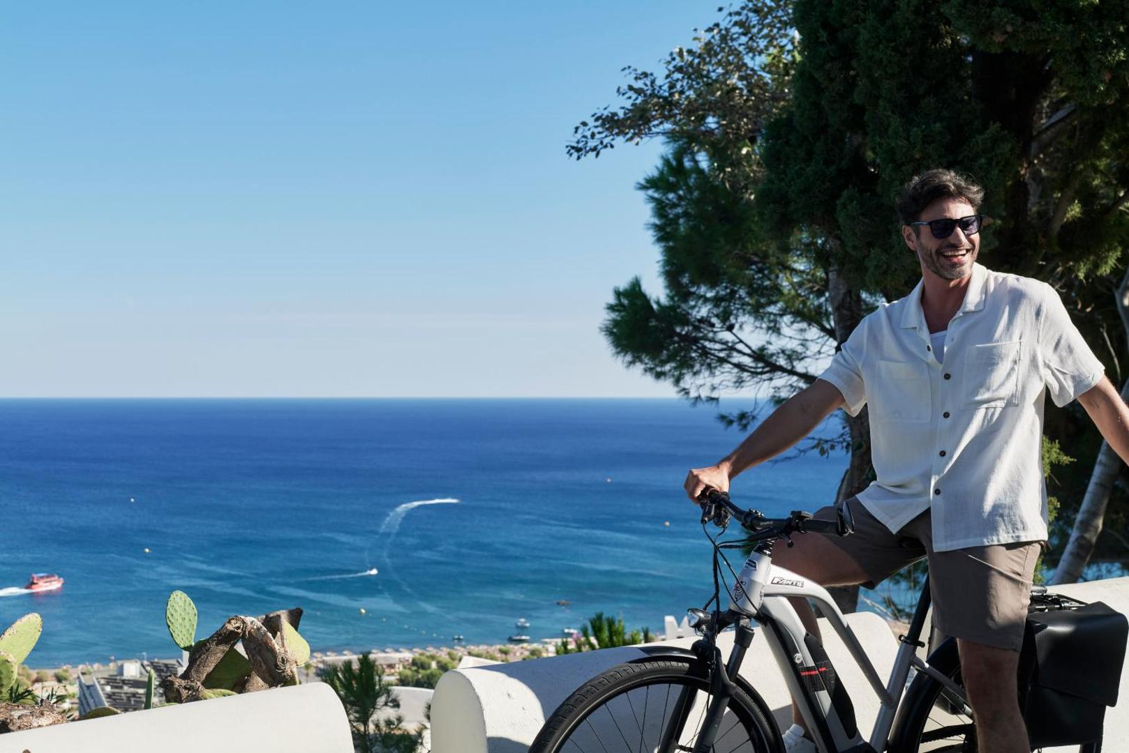 Vīrietis ar velosipēdu smaida uz kalna fona ar skatu uz jūras piekrasti.