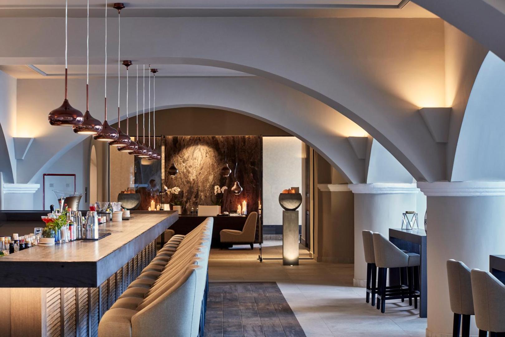 Elegants viesnīcas bārs ar gariem krēsliem, modernām lampām un arkveida griestiem.