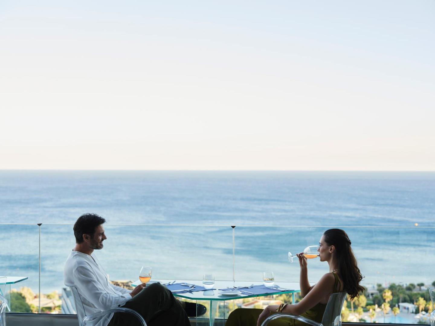 Pāris bauda dzērienus pie stikla galdiņa uz terases ar skatu uz zilajiem ūdeņiem.