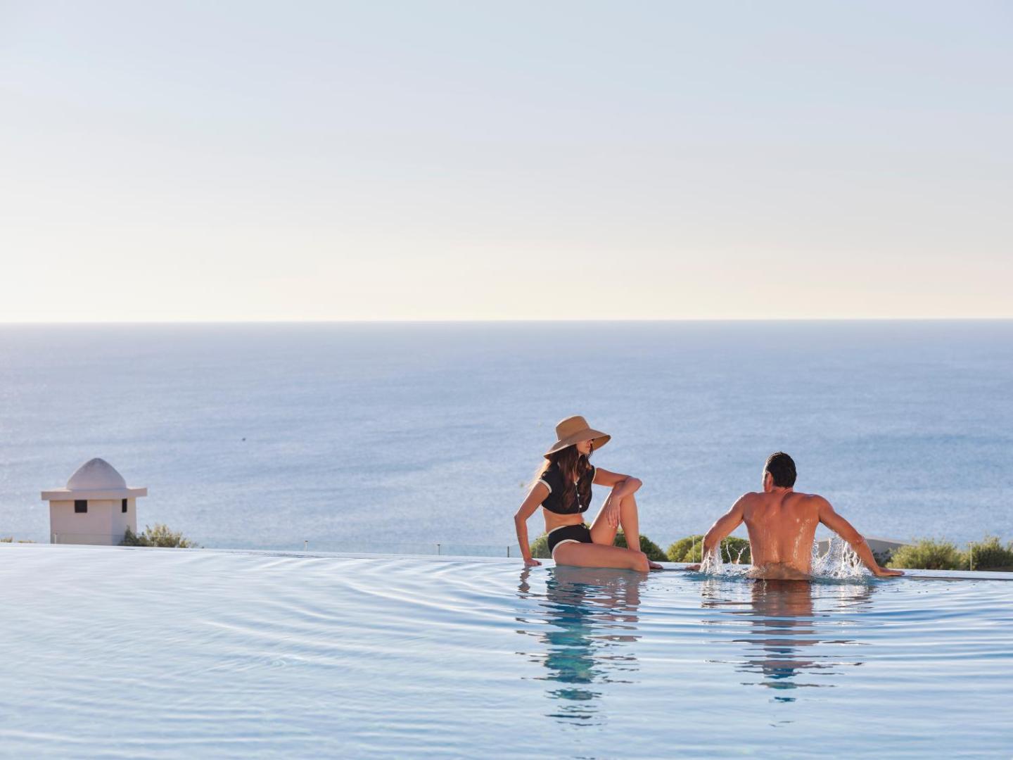 Pāris atpūšas bezgalības baseina malā, vērojot saulrietu virs jūras.