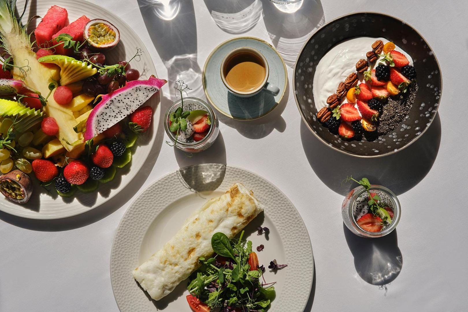 Bagātīgas brokastis ar svaigiem augļiem, jogurtu un kafiju uz balta galda.