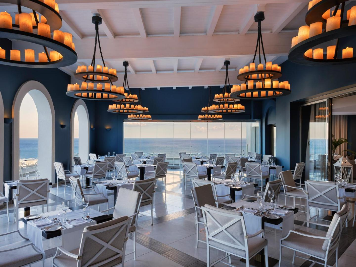 Eleganta viesnīcas restorāna zāle ar zilām sienām, greznām lustrām un skatu uz jūru.