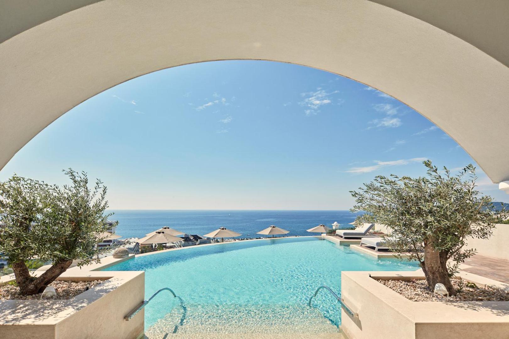 Bezgalības baseins ar skatu uz jūru, ierāmēts ar baltu arku un olīvkokiem.