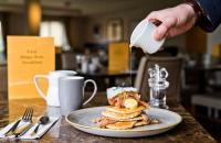 a person pouring syrup onto a plate of pancakes at voco Fiorello - LaGuardia Airport by IHG in Queens