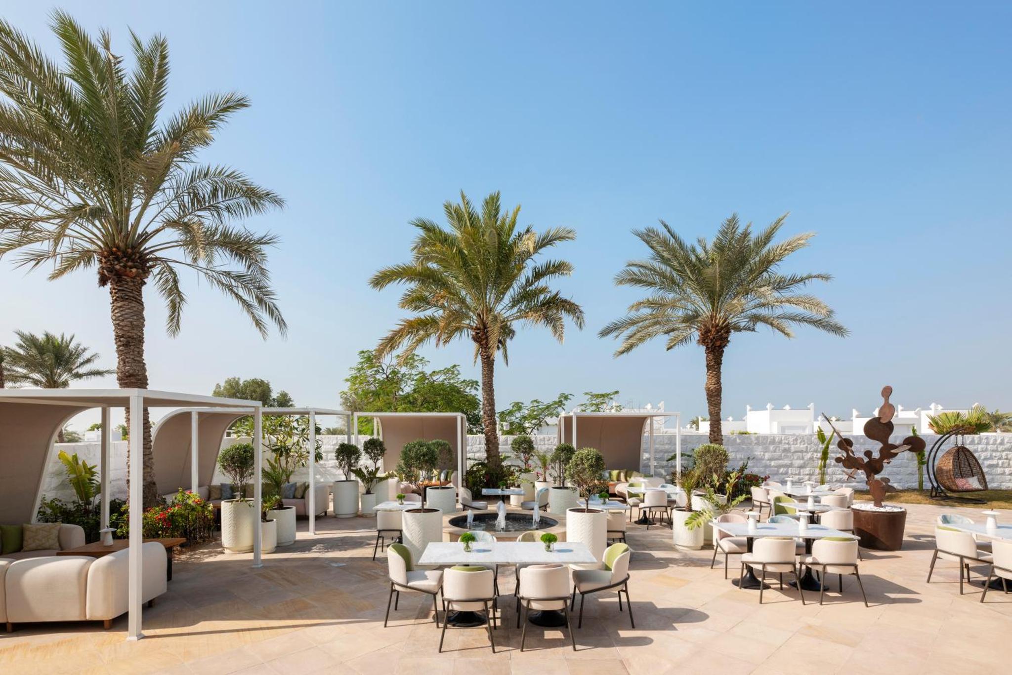 a hotel patio with tables and chairs and palm trees at Raffles Al Areen Palace Bahrain in Manama