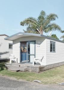 a small white house with two chairs and a palm tree at Mercury Bay Holiday Park in Whitianga