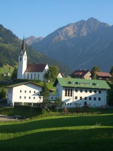 ein großes weißes Gebäude mit einer Kirche mit Bergen im Hintergrund in der Unterkunft Herzen´s Landhaus in Hirschegg