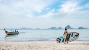 un uomo che cammina sulla spiaggia con una tavola da surf e una barca di The Tubkaak Krabi Boutique Resort - SHA Extra Plus a Tab Kaek Beach