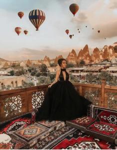 una mujer con un vestido negro sentada en un balcón con globos de aire caliente en Goreme Cave Rooms&Spa, en Göreme