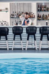 a man standing behind a bar with three chairs next to a pool at Mykonos Princess Hotel in Agios Stefanos