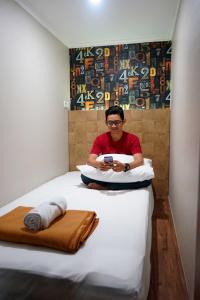 un hombre sentado en una cama mirando su celular en FixOn Capsule Hotel, en Purwokerto
