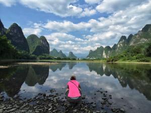 a woman sitting on the shore of a river at Yangshuo Yunshe Mountain Guesthouse in Yangshuo
