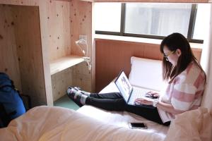 a woman sitting on a bed with a laptop at Mezame Hostel in Nagasaki