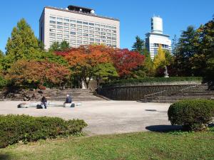 two people sitting on benches in a park with a large building at Koyo Grand Hotel in Sendai