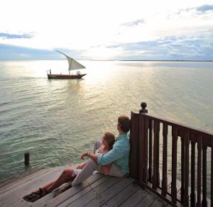 a couple sitting on a dock with a boat in the water at The Residence Zanzibar in Kizimkazi