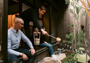 Zwei Menschen sitzen auf dem Felsvorsprung eines Hauses mit einem Mann, der einen Holzlöffel hält. in der Unterkunft Inari Ohan in Kyoto