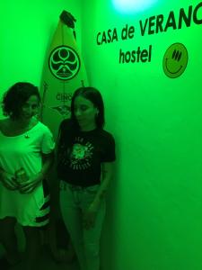 two women standing in front of a green wall with a surfboard at Pinamar Hostel Casa de Verano in Pinamar