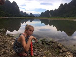 a young girl sitting on the shore of a river at Yangshuo Yunshe Mountain Guesthouse in Yangshuo