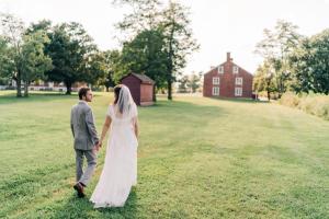 a bride and groom walking through a field with a barn at Shaker Village of Pleasant Hill in Harrodsburg