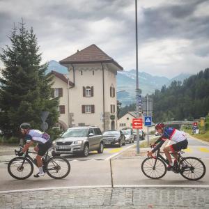 two men riding bikes down a street with a building at Sust Lodge am Gotthard in Hospental