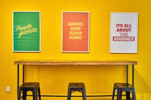 three posters on a yellow wall with three stools at Hostal Americana in Guadalajara
