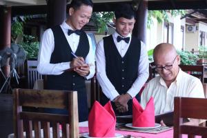 two men in tuxedos standing next to a table with a man cutting at Pon Arena Hotel in Muang Không