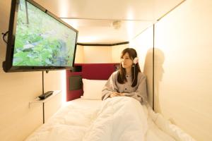 Una donna seduta su un letto di fronte a una TV di Matsue Urban Hotel CubicRoom a Matsue
