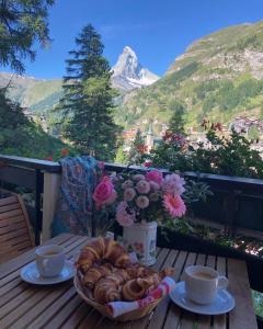 einen Tisch mit einer Schüssel Croissants und Tassen sowie einem Korb Brot in der Unterkunft Haus Oasis in Zermatt