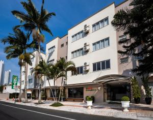 a tall white building with palm trees in front of it at Hotel Rosenbrock in Balneário Camboriú