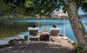 un homme et une femme assis sur des chaises sous un parasol sur un lac dans l'établissement Appart'Hôtel LIDO au bord de l'eau, à Gérardmer 24 autres photos