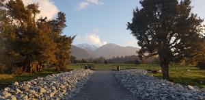 a gravel road with trees and mountains in the background at Cosy Cabin in the Paddocks - Breakfast Included in Franz Josef