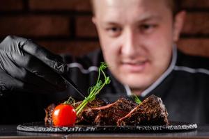 Een man in zwarte handschoenen die een steak eet met een tomaat. bij Deluxe Hotel Kupava in Lviv