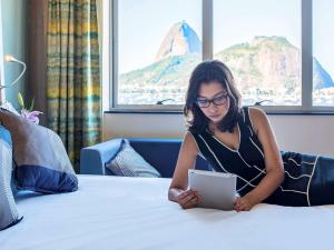 una donna seduta su un letto usando un tablet di Novotel RJ Praia de Botafogo a Rio de Janeiro