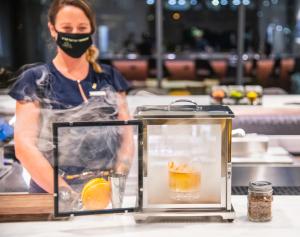 Uma mulher com uma máscara facial atrás de duas bebidas. em Hotel Eleo at the University of Florida em Gainesville