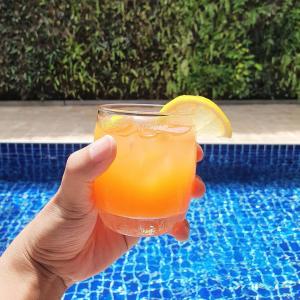 a hand holding a glass of orange juice next to a pool at Dinsomon Hotel in Bangkok