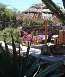 a patio with a thatch umbrella and some flowers at Le casette di Marilena Agave Iucca in Pantelleria