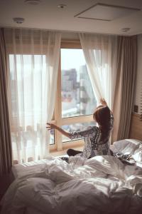 una mujer sentada en la cama mirando por una ventana en Kenny Stay Jeju Seogwipo, en Seogwipo