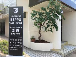 un árbol delante de un edificio con un cartel en J-Hoppers Beppu Guesthouse ジェイホッパーズ別府ゲストハウス, en Beppu