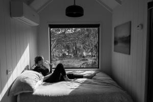 a man sitting on a bed looking out a window at Big Yard Escapes - Two Off-Grid Tiny Houses on the Mowamba River in Jindabyne