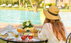 Una mujer con sombrero sentada en una mesa con comida junto a la piscina en Les Jardins De Zyriab, en Aït Hamid