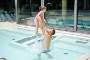 a young girl standing on a mans back in a swimming pool at Alpholiday Dolomiti Wellness & Family Hotel in Dimaro