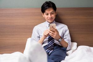 un hombre sentado en la cama mirando su celular en Comfort Hotel Narita, en Narita
