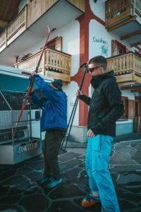 dwóch mężczyzn ze sprzętem narciarskim stojących przed budynkiem w obiekcie Casa di Franz w mieście Vigo di Fassa