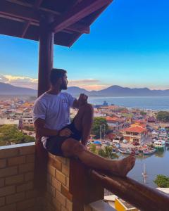 Ein Mann, der auf einem Felsvorsprung sitzt und über eine Stadt blickt. in der Unterkunft Hostel Vista da Barra in Florianópolis