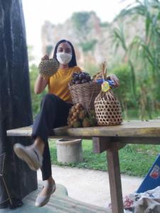 une femme portant un masque assise sur une table avec une corbeille de fruits dans l'établissement โฮมสเตย์บ้านนายสิงโตเขาคูหา, à Ban Khu Ha Nai (1)