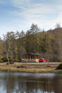 a cabin by a body of water with a red car at 2-Bed Cottage with Hot Tub at Loch Achilty NC500 in Strathpeffer