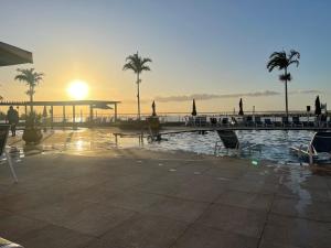 uma piscina com palmeiras e um pôr-do-sol ao fundo em Golden Lake Apart Hotel Arraial do Cabo em Arraial do Cabo