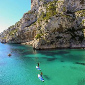 two people riding surfboards in the water near a mountain at A 500 mètres des plages et du port du brusc, Les terrasses des embiez avec piscine et spa in Six-Fours-les-Plages