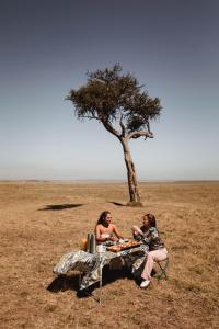 two women sitting at a table in a field with a tree at Emboo River Camp in Talek