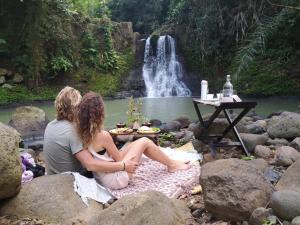 dos mujeres sentadas en las rocas frente a una cascada en Galalima Glamping en Tabanan